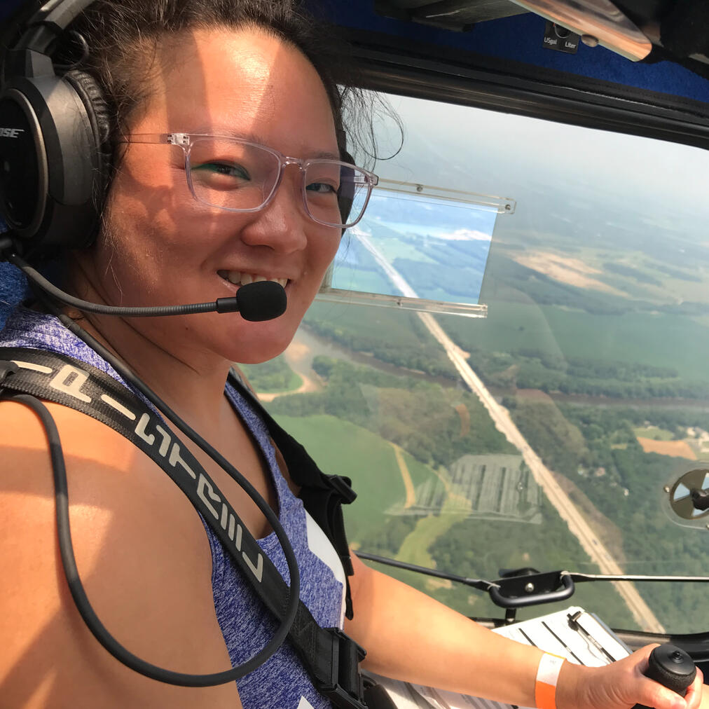 Sheila Xu as a Pilot Sheila, an Asian-American woman in her 30s, smiles at the camera while wearing glasses and a headset, piloting a plane with a scenic view of Indiana's green plains visible through the window behind her.