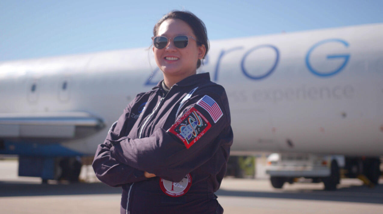 Sheia in Her Zero-G Flightsuit Sheila is in a dark navy-colored flight suit with mission patches and wearing her sunglasses, stands in front of a Zero G aircraft on a sunny day.