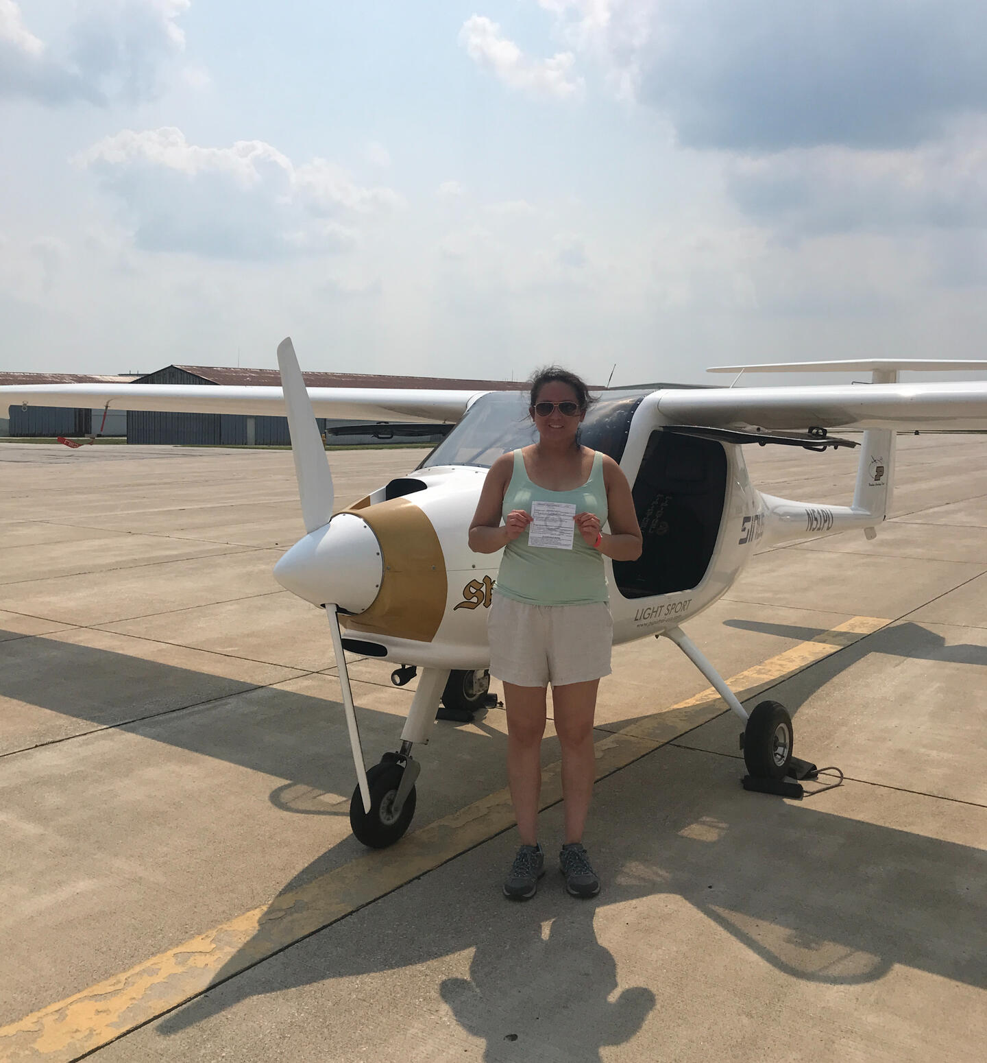 Sheila Gets Her Pilot Certificate! Sheila stands in front of a small white, 2 seater Pipistrel airplane on an airport tarmac, holding her new flight certificate, under a partly cloudy sky.