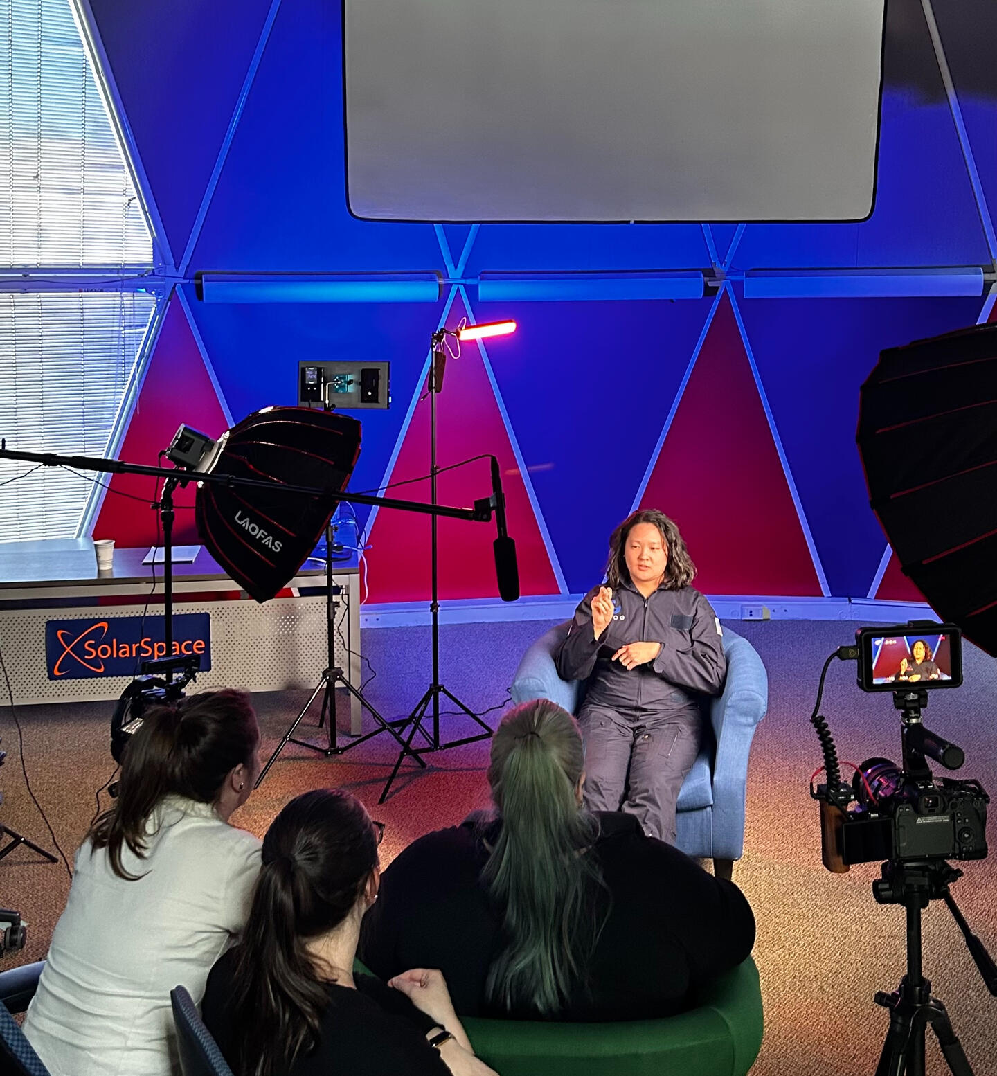 Interviewing Sheila at BioSphere2 Sheila sits in a blue chair speaking on camera during a recorded interview, with three people watching, studio lights, and filming equipment set up in a geodesic dome at BioSphere2 facility in Tucson, AZ.
