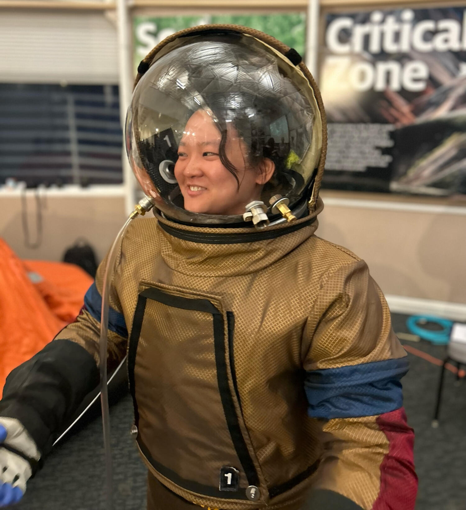 Sheila in Her Spacesuit! Sheila smiles while wearing a bulky brown spacesuit with a clear helmet, standing indoors in front of informational posters at BioSphere2 in Tucson, Arizona.